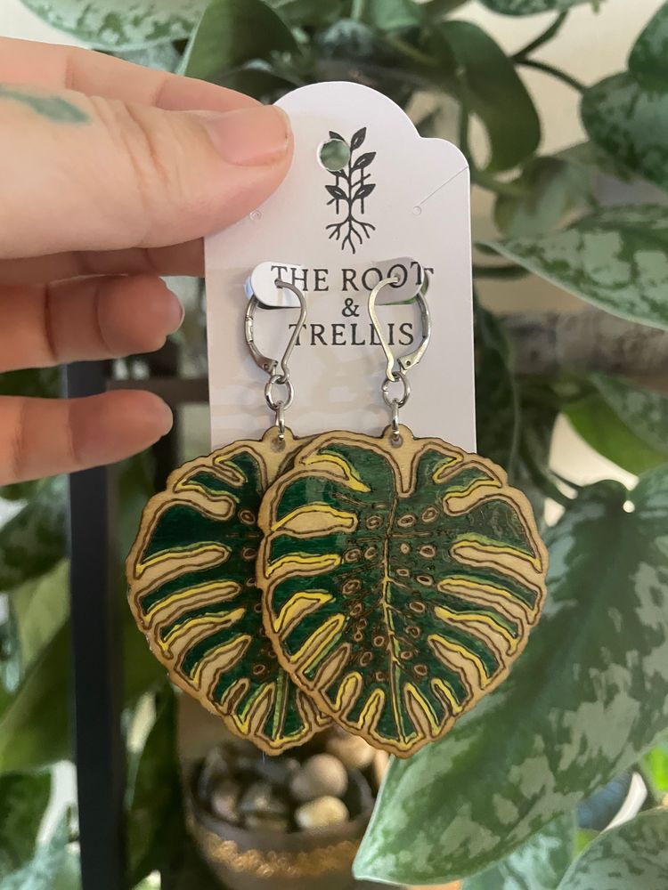 An image of a pair of green monstera leaf earrings with a mature fenestrated leaf design, being held by the hand of a person in front of a botanical backdrop.