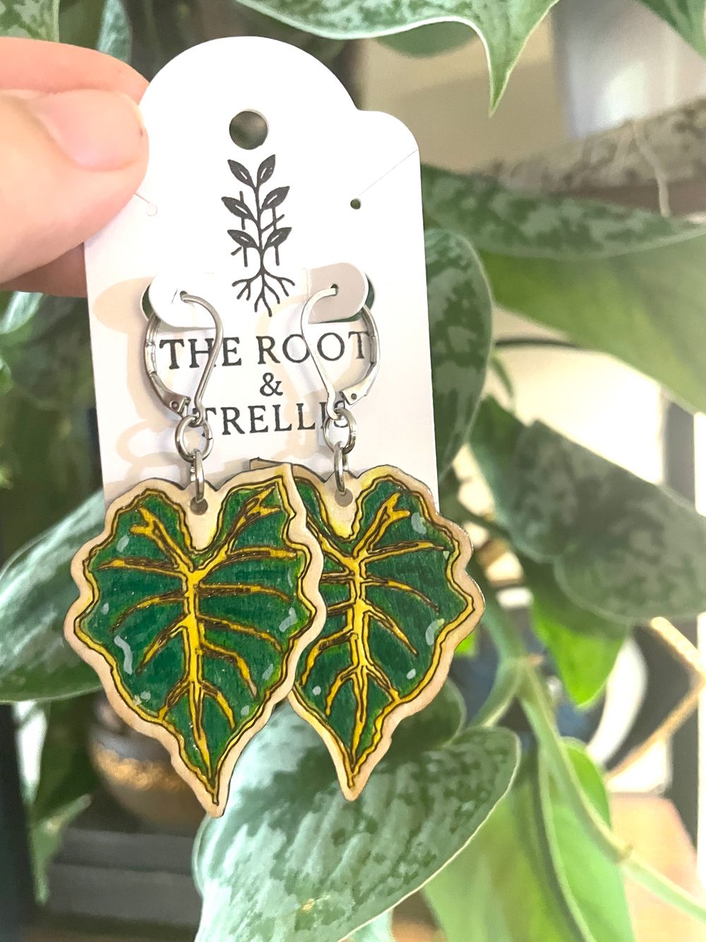 An image of a pair of green alocasia-leaf earrings being held by the hand of a person in front of a botanical backdrop.